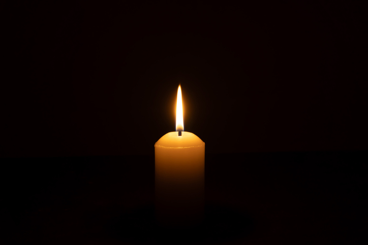 A single burning candle flame or light glowing on a big white candle isolated on black or dark background on table in church for Christmas, funeral or memorial service