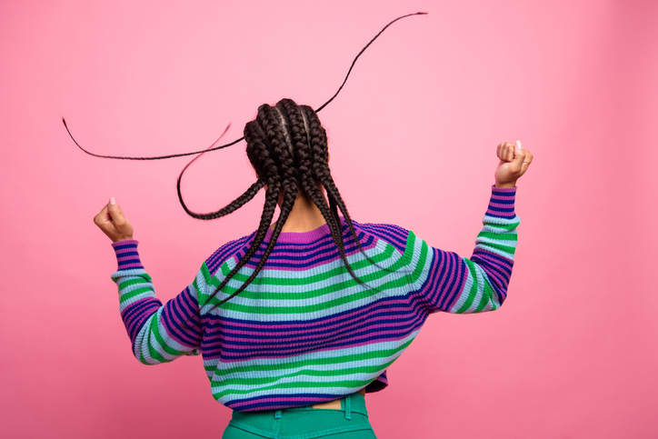 Joyful young woman dancing with braids, colorful striped sweater against pink background, expressing carefree energy and happiness.