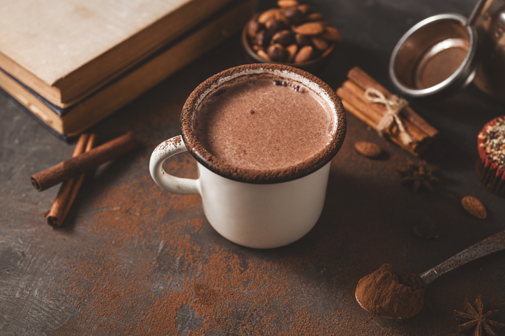 Hot chocolate in white enamel mug on dark rustic surface with nuts, cocoa powder, cinnamon sticks and cupcake, seasonal cozy winter drink concept