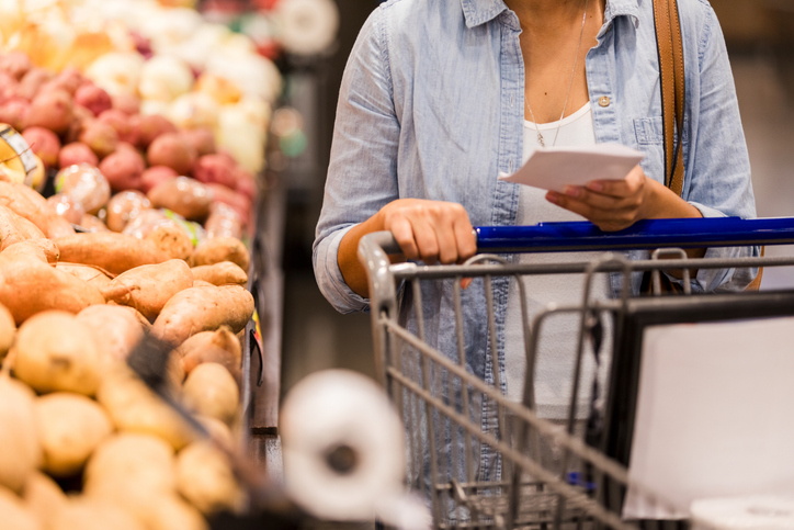 Woman checking shopping list while pushing cart past produce in supermarket aisle.